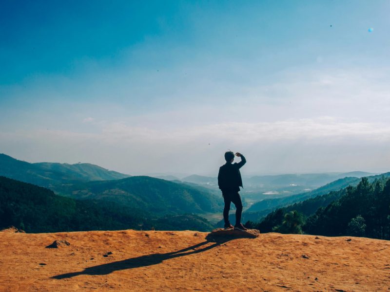A lone traveler stands on a hilltop, gazing over a vast mountain landscape under a clear blue sky.