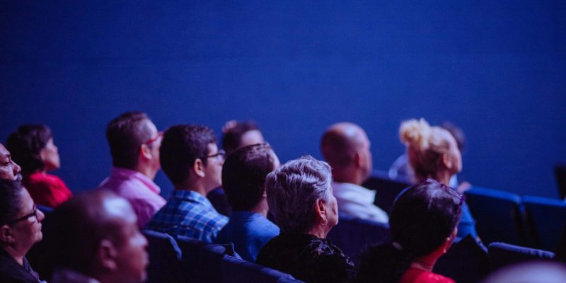 An attentive group of adults seated at an indoor conference, focusing on a presentation.