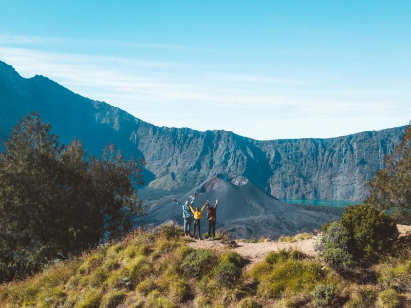 Three hikers raise their arms in joy at the crater rim of Mount Rinjani, Lombok, Indonesia.