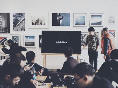 A group of adults attending a casual business presentation in a modern art space, featuring a TV display and framed art.